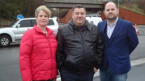 Councillors Steph Exell, Kevin Small and Jim Robbins. Steph has light hair which is tied back. She is wearing a red coat. Kevin Small has short dark hair and is wearing a black coat. Jim Robbins is bald with facial hair. He is wearing a blue blazer and jeans. The three of them are stood in a line side by side, looking at the camera and smiling. 
