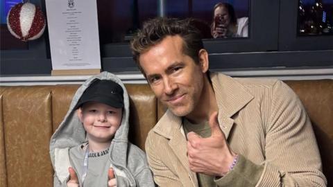 Riley Bains and Ryan Reynolds sit together on a coach, smiling at the camera. Nine-year-old Riley is wearing a grey hoodie and a black cap. Reynolds has short brown hair and is wearing a light brown jacket.