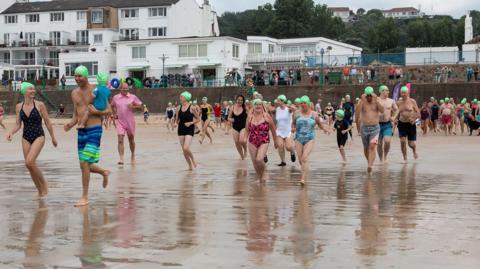 There are dozens of men, women and children wearing swimsuits and green swimming hats running across the beach to the sea. They are smiling and laughing. It is a dull day. There are spectators lined up on railings above the sea wall and houses behind them.