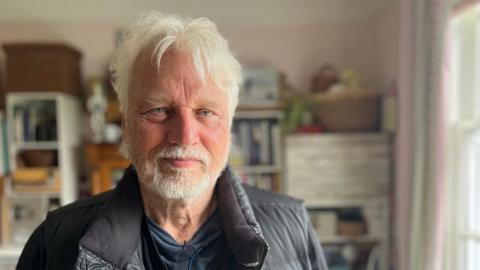 David Knopfler, wearing a black gilet, with a blurred background of shelves at his home