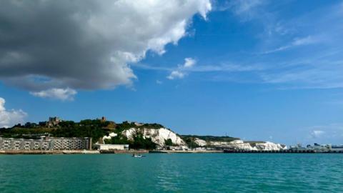 White cliffs on a seashore meet turquoise water under a dark grey cloud with clear blue sky surrounding it.
