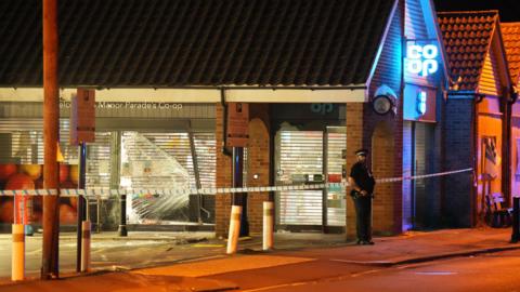 An exterior view of a parade of shops in Worthing that have been ram raided by a vehicle. The metal shutter of a supermarket has been smashed in, as has the glass frontage of the store, while a concrete bollard is at an angle. A lone police officer stands guard at the scene.