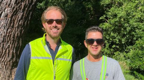 Ben Rhodes on the left is dressed in a high viz jacket with sunglasses, blond slicked back hair and a beard. Michael Ellis is seen on the right in a grey t-shirt, high viz straps over his shoulders, bulky sunglasses and dark, short hair.