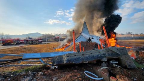 A large piece of debris from a plane lies on the ground next to a runway and is on fire