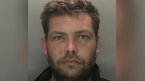 Custody image of Joshua Allen. He has a flushed face, beard and light moustache. He has a thick crop of styled brown hair. His black clothing is only visible under his neckline.