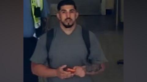 A man in a grey V-neck t-shrt hold a phone in both hands as he walks on a tiled floor in this CCTV still.