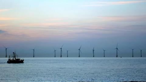 Wind turbines in the sea on the horizon, a trawler is moving from left to right in the foreground.