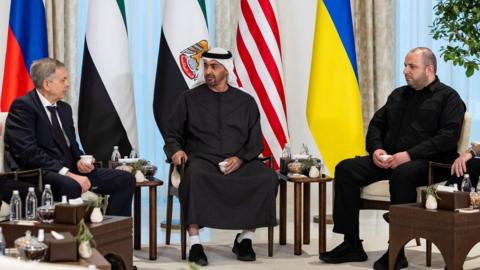 A handout photo released by the UAE Presidential Court shows UAE President Mohamed bin Zayed Al Nahyan (centre), meeting Rustem Umerov, Secretary of the National Security and Defense Council of Ukraine (R), Igor Kostyukov, Chief of the Main Directorate of the General Staff of the Russian Armed Forces (L)during trilateral talks between the United States, Russia and Ukraine at Al Shati Palace in Abu Dhabi. The participants are sitting in a semi-circle with the Russian, UAE, US and Ukrainian flags behind them.