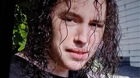 Vlad Nikolin-Caisley looks at the camera through his long, curly and black hair which hangs down to his shoulders. He is wearing a black t-shirt.