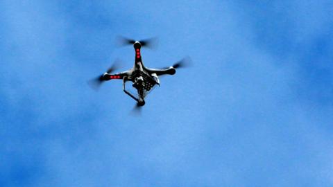 A grey drone with four propellers and red lights on the underside flies across a blue sky