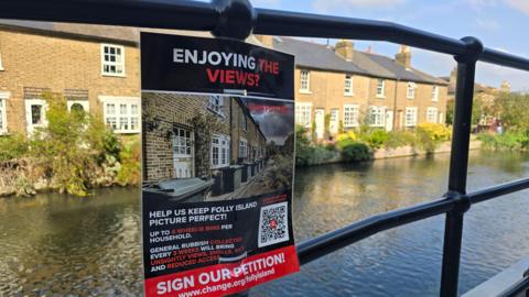 A view of a row of terraced houses alongside a river, a sign warning about the new bins is pinned to the railing