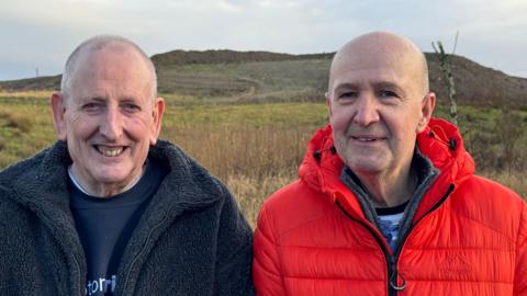 An image of Trevor Turner and Trevor Lawson. Trevor Turner is wearing a navy fleece jacket with a blue jumper, Trevor Turner is wearing a red puffer jacket, both are smiling. They are standing with the landfill site in the background.