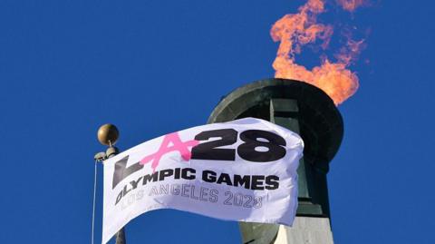A white flag reading "LA 28 Olympic Games Los Angeles 2028" is seen waving in front of a lit cauldron. The Olympic flame is towering overhead 