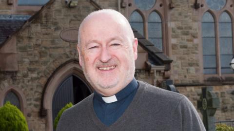 Fr John Murray stands outside in front of a stone-brick church. He wears a grey jumper over a navy shirt with a white priest collar. 