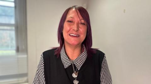 Claire Curran is smiling. Her shoulder length hair is styled in maroon. She's wearing a black vest jumper with a black and white blouse underneath and heart shaped pendants are around her neck. She's standing in a room. 