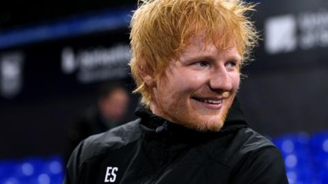 Ed Sheeran sits in Portman Road stadium. He looks away from the camera while smiling. He has long red hair and a red beard. He wears a black rain coat.
