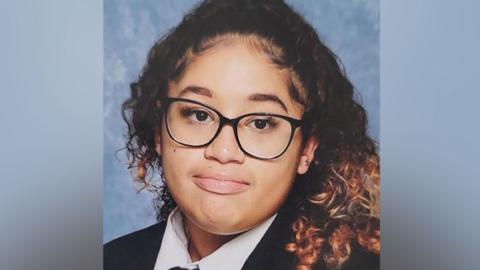 A girl with long curly hair, wearing glasses and a school uniform