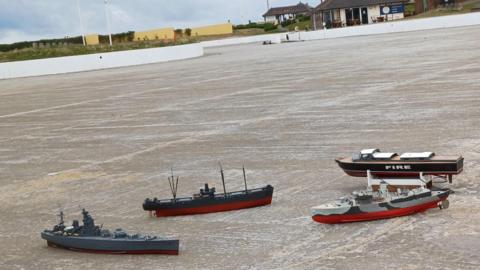 Four model boats laying on the concrete of the empty boating lake in Coatham, Redcar. The structure has a curving shape and is near the sea. The Boathouse and Terrace cafe is visible in the background.