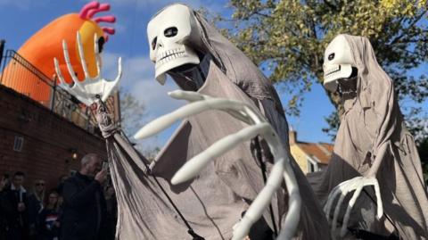 Two skeleton figures walking through the streets of Lincoln. They are wearing grey capes. An orange inflatable monster is in the back left of the photo. 