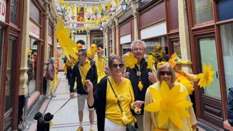 People dressed in yellow smile and wave yellow flowers and wear yellow clothing as they walk through the indoor market in Hull.