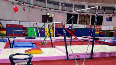 Gym equipment installed at the gym club in Jersey. A number of gymnastic bars have been installed. White and pink mats are on the floor of the gym. A red balloon is floating in the air. 