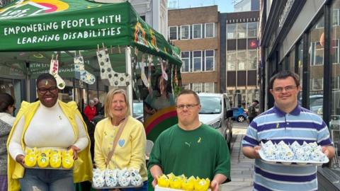 A group of four people stand in front of a green market stall tent labelled “Les Amis – Empowering People with Learning Disabilities.” They are smiling and holding trays of decorated items shaped like small chicks and sheep. The scene takes place on a city street with other people browsing the stall in the background.
