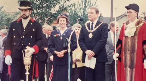 Four people standing in front of a microphone at a ceremony. The woman and man in the centre are wearing formal attire with the man donning a mayoral chain. The two men on either side are wearing ceremonial outfits.