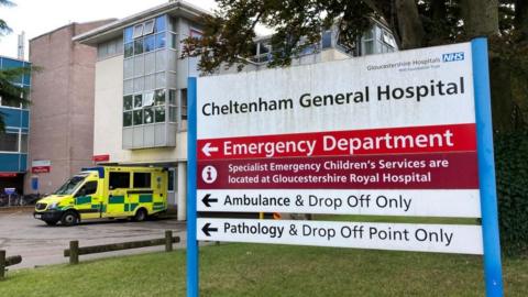 The sign for Cheltenham General Hospital's Emergency Department on a grass lawn in front on the main building, next to a parked ambulance
