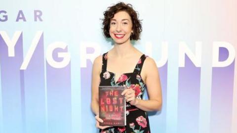 Author Andrea Bartz at an event holding a hardback copy of her novel The Lost Night. She is wearing a black sleeveless dress with a rose pattern  