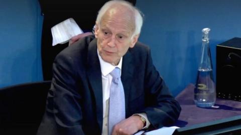 The chair of the inquiry, wearing a blue suit and tie, sits with his arm leaning on a desk