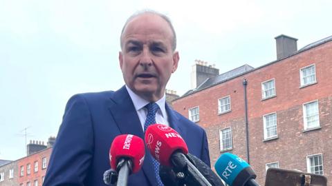 Micheál Martin, a man with short, receding grey hair, speaks into microphones while standing on a street in central Dublin. He is wearing a navy blue suit and a navy blue polka dot tie. A terrace of tall, red brick Georgian town houses line the road behind him. 