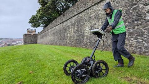 A man pushing a ground penetrating radar scanner over an area of grass in Derry close to the city's walls which can be seen in the background