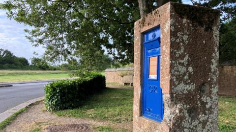 A blue post box in a concrete blocks which has dents and chips from shrapnel damage from World War Two. Behind it are a hedge, wall, trees and a road.