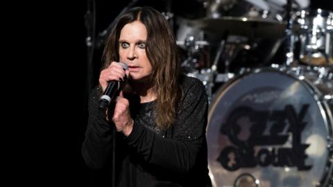 Ozzy Osbourne, in a black outfit, is on stage holding a microphone. He is stood in front of a drumkit which features a logo with his name on it.