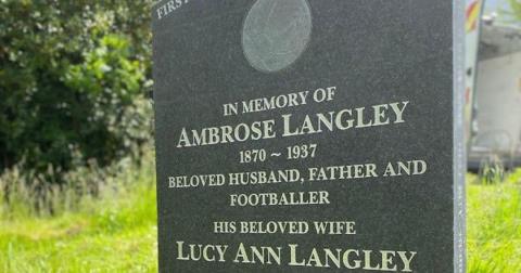 A close-up of a dark grey granite headstone with the wording, 'In memory of Ambrose Langley 1870 - 1937 beloved husband, father and footballer, his beloved wife Lucy Ann Langley'