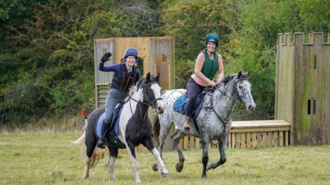 Two women on horseback smiling, one waving, as they ride past the camera. 