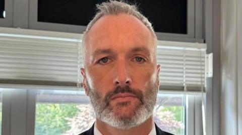 Det Con Lee Harshey-Jones. He looks at the camera with a neutral expression, and wears a white shirt, black suit jacket and bronze tie. He has short grey hair and short stubble.