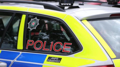 Close up of the rear passenger side of a yellow and blue coloured police car with Battenberg markings with tinted windows at the rear. One of the windows has 'police' written in red on it alongside the PSNI badge.