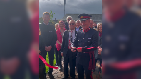 A man in a military uniform cuts a red ribbon in front of a crowd of smiling people