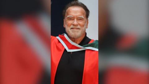 Arnold Schwarzenegger smiles at the camera. He has short, dark hair and a short, white beard. He wears a red graduation gown.