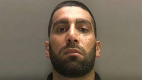 Mugshot of a man with a black beard and hair with a scar on his nose