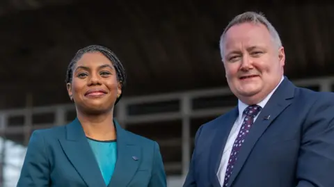 Kemi Badenoch and Darren Millar stood next to each other. Badenoch is stood to the left and is slightly shorter than Millar. Both are smiling to the camera.