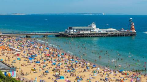 Bournemouth beach, a sandy beach which is packed with hundreds of people, tents and umbrellas. Bournemouth pier is in the background.