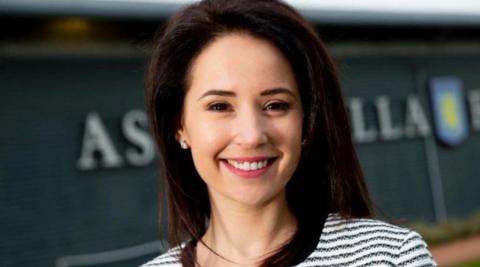 A woman with dark hair and a black and white top is smiling at the camera. The Aston Villa club crest is blurred in the background.