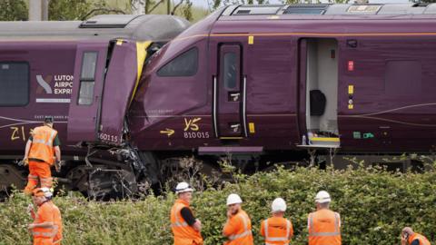 Two trains are seen crashed together with people wearing orange protective clothing standing alongside.