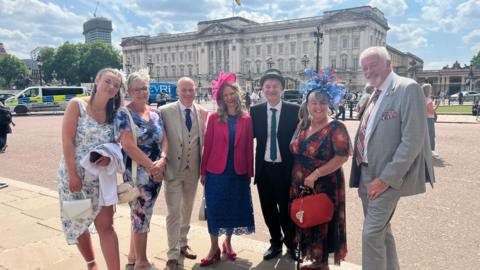 Left to Right: Emma Sellars, Ms Sellars' mother, Stuart Wills, Mr Wills' partner Karen, Scott Dalton, Liz Quiney and Ms Quiney's partner. They are all dressed in smart clothes and are stood in front of Buckingham Palace.