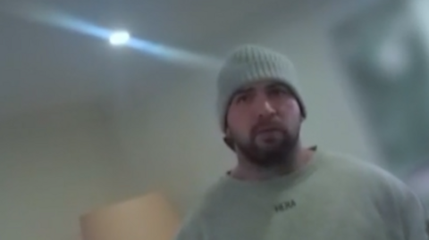 Bodycam footage of a man in a grey sweather and grey beanie hat stands in his living room with a lamp and a television behind him