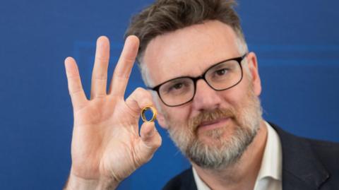 Charles Hanson with short brown hair, a brown and grey beard and glasses holds a gold ring between the thumb and index finger of his right hand