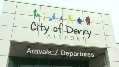 The front of City of Derry airport - it has arrivals and departures sign and above the sign, it shows a number of drawings representing the people who will use the airport. These include a wheelchair user, a couple, a single backpacker, a child, a businesswoman and a person with a bunch of flowers.