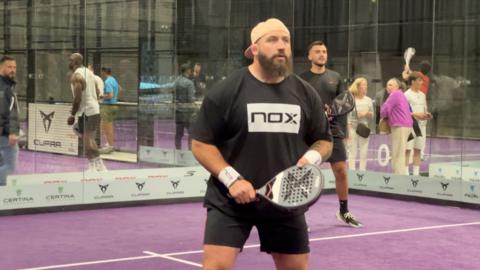 A man in a black shirt with a white box on it, holding a padel racket.